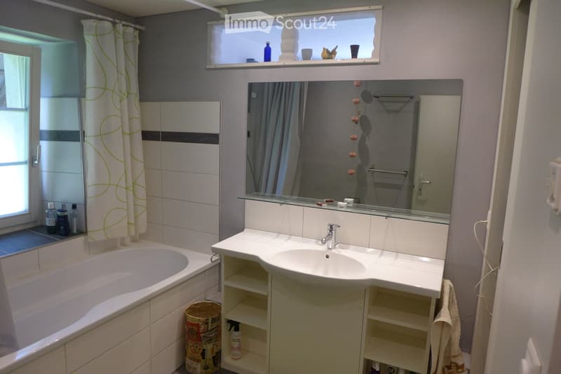 Bathroom with a bathtub, sink, mirror, and cabinet. The room features a window with a curtain, a shelf above the window holding cups and bottles, and a towel hanging on the side.