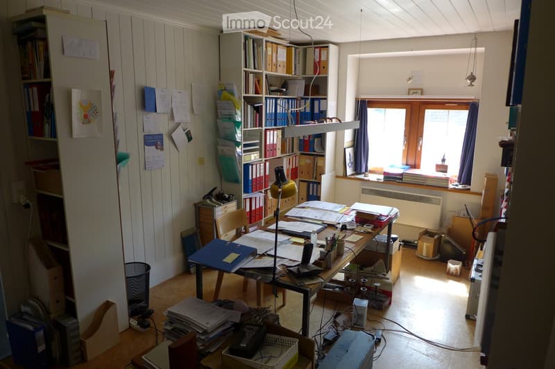 Office space with a cluttered desk, bookshelves, filing cabinets, and a window with curtains.