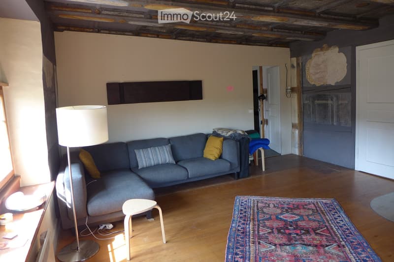 Living room with wooden flooring, grey sectional sofa, colorful throw pillows, floor lamp, small white stool, and a patterned rug.