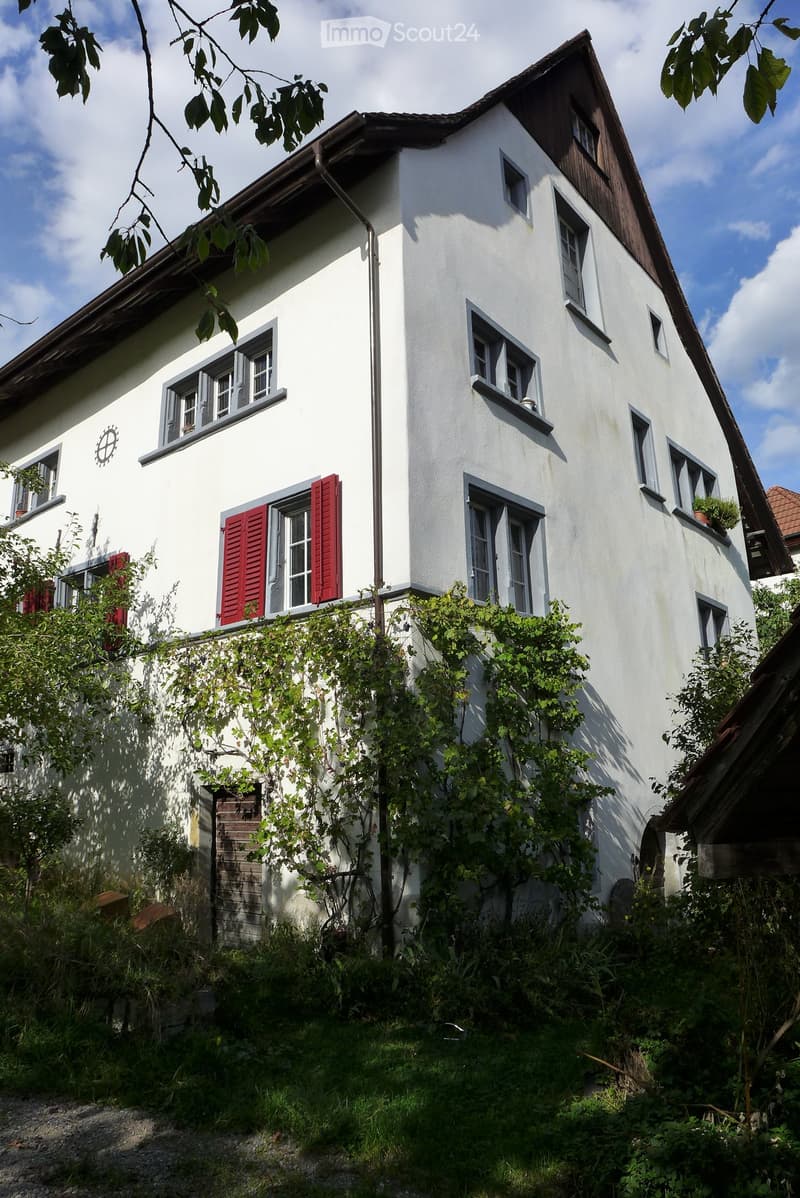 Two-story white house with grey-framed windows, red shutters, and a sloped roof. Ivy covers part of the lower wall, and the surrounding area is overgrown with vegetation.