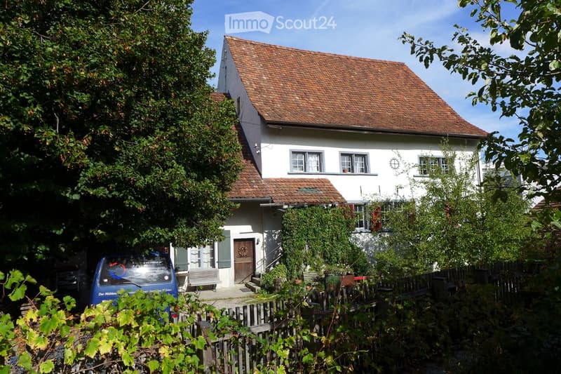 Two-story white house with red-tiled roof, multiple windows, and a small porch. A blue van is parked nearby, and the house is surrounded by greenery and a wooden fence.