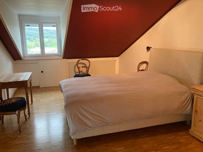 Cozy attic bedroom with wooden flooring, slanted ceiling, large windows offering a scenic view, a neatly made bed, a wooden table, and two chairs.