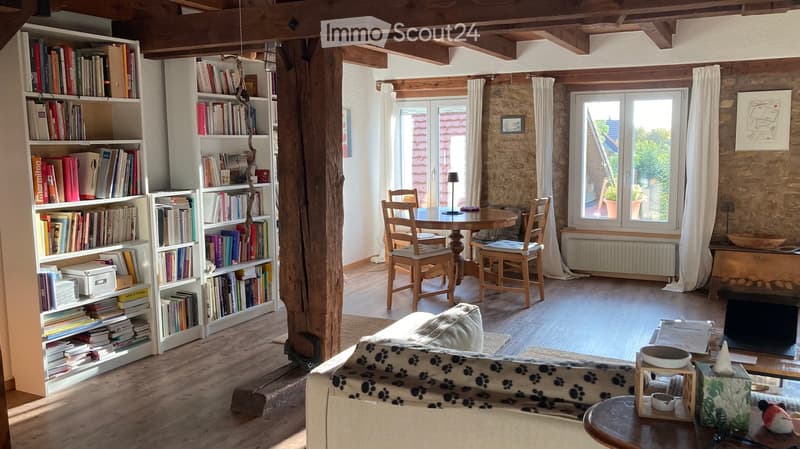 Cozy living room with wooden beams, exposed stone wall, large bookshelf, sofa, dining table, and chairs.