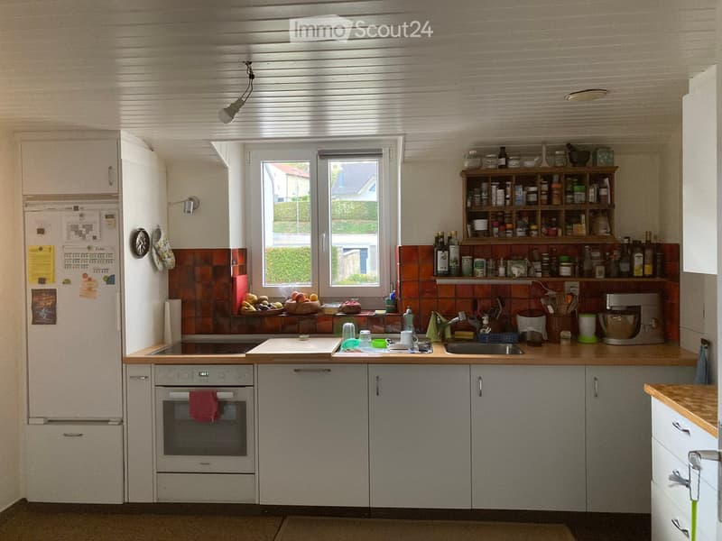 The kitchen features a refrigerator, oven, sink, and various appliances and utensils. The countertops are wooden, and there are cabinets and shelves with items like bottles and bowls.