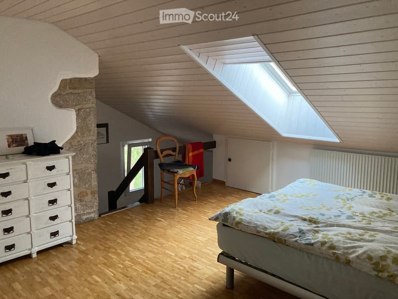 Bright attic bedroom with wooden floors, slanted wooden ceiling, skylight windows, bed with floral bedding, wooden chair, dresser, radiator, and a small window with a view.