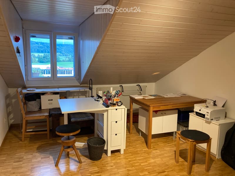 Well-lit attic workspace with wooden floors, slanted wooden ceiling, large windows offering a scenic view, two desks with chairs, a printer, and various office supplies.
