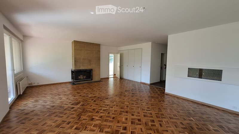 Empty living room with wooden parquet flooring, white walls, fireplace, large window, and built-in white closet.
