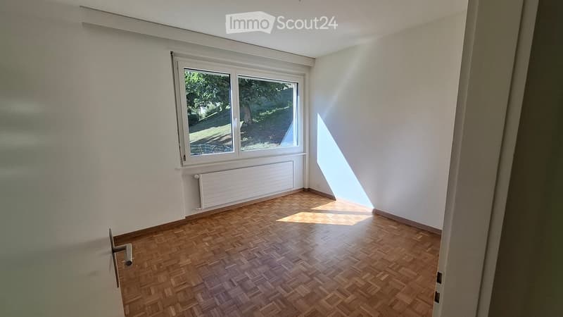 Empty room with wooden herringbone flooring, white walls, and a large window with a view of greenery outside. The room has a radiator below the window and a door visible on the left side.