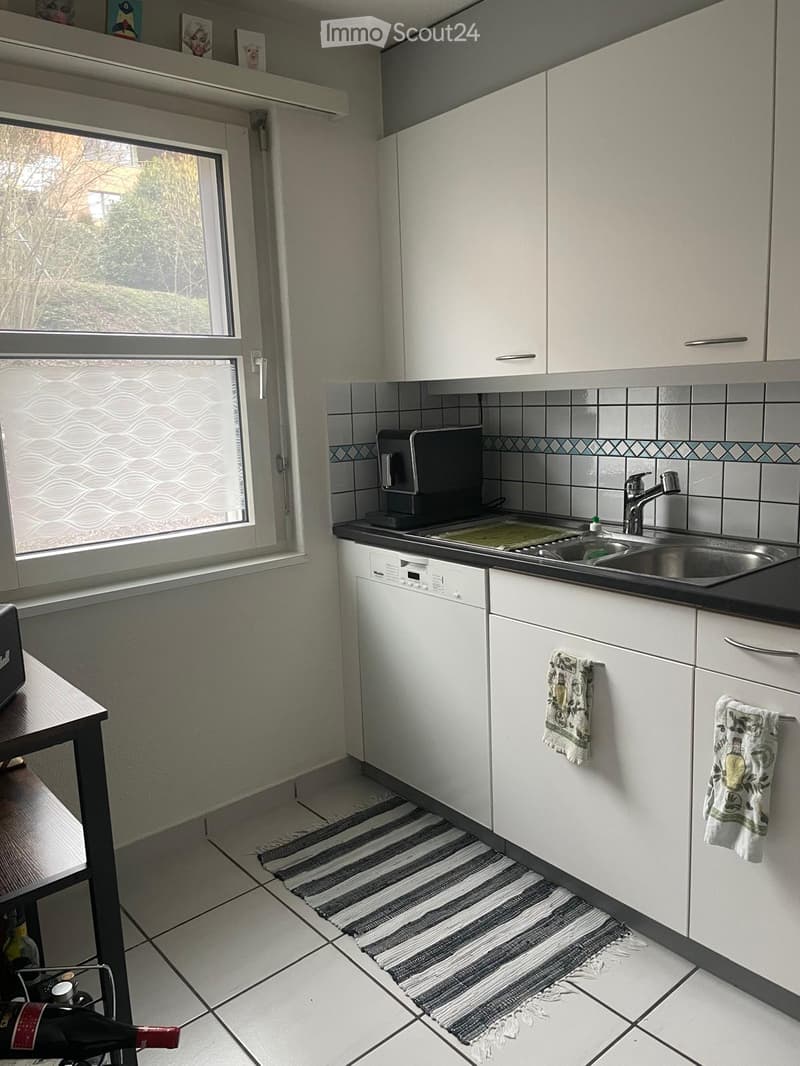 Modern kitchen with white cabinets, tiled backsplash, stainless steel sink, dishwasher, coffee maker, and a small table with a wine rack.