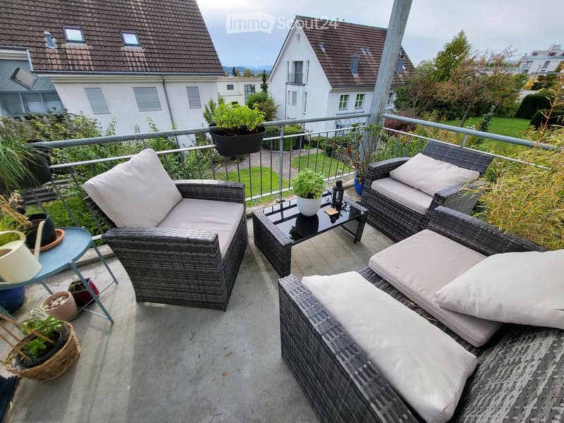 A well-furnished balcony with wicker chairs, a sofa, a coffee table, and several potted plants, overlooking a residential area.
