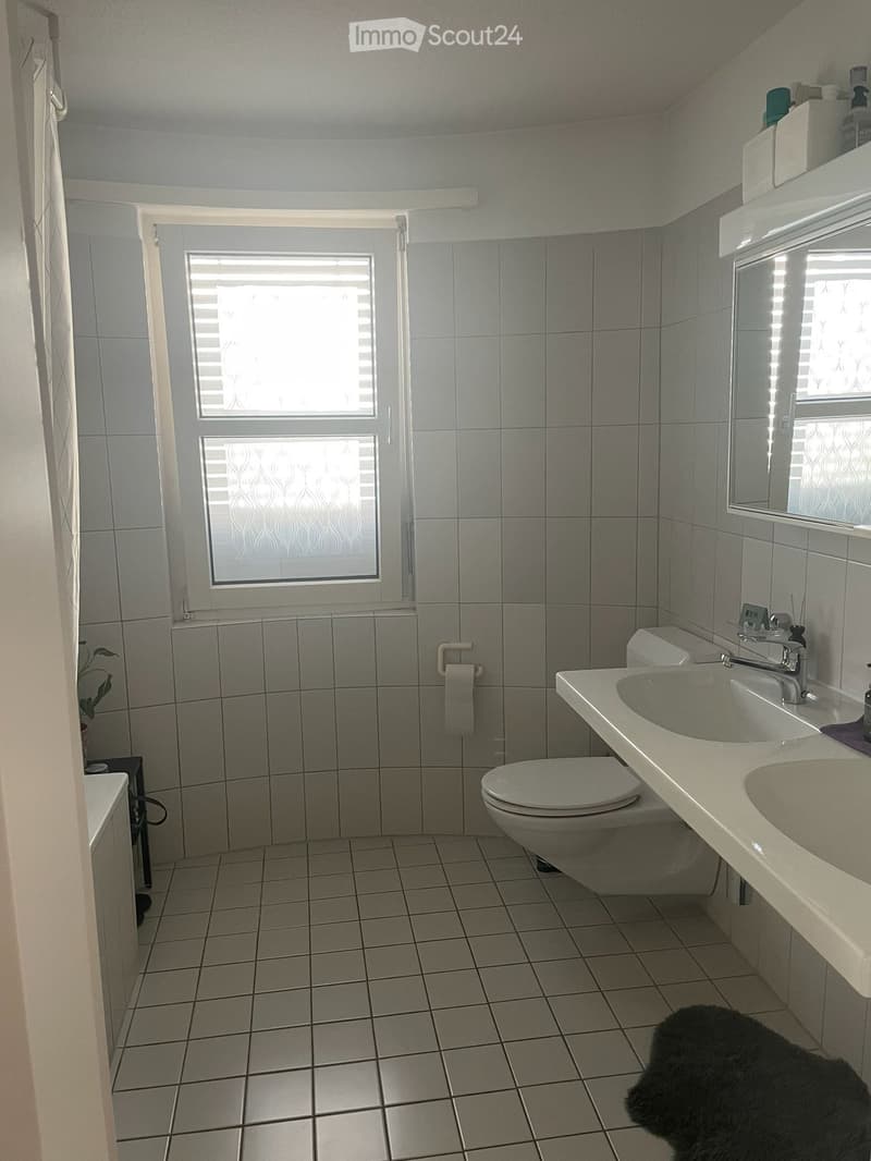 A modern bathroom with a toilet, double sink, mirror, and tiled walls and floor. The room features a window with blinds and a small black rug on the floor.