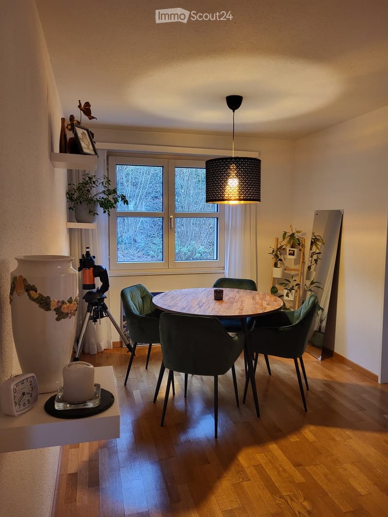 Dining area with a round wooden table surrounded by green chairs, a large vase, a tripod, a clock, and a mirror. The room has wooden flooring and large windows with a view of trees.
