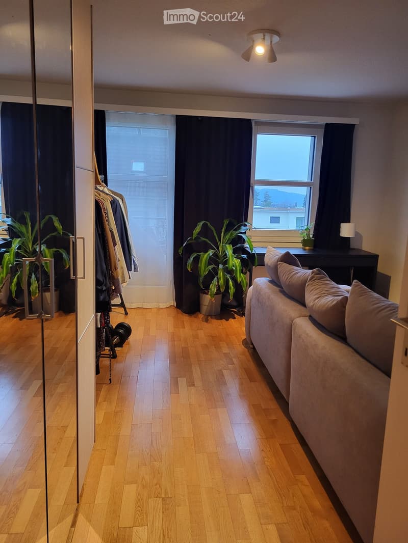 A modern living room with wooden flooring, a grey sofa, potted plants, a small table, and a wardrobe with hanging clothes.