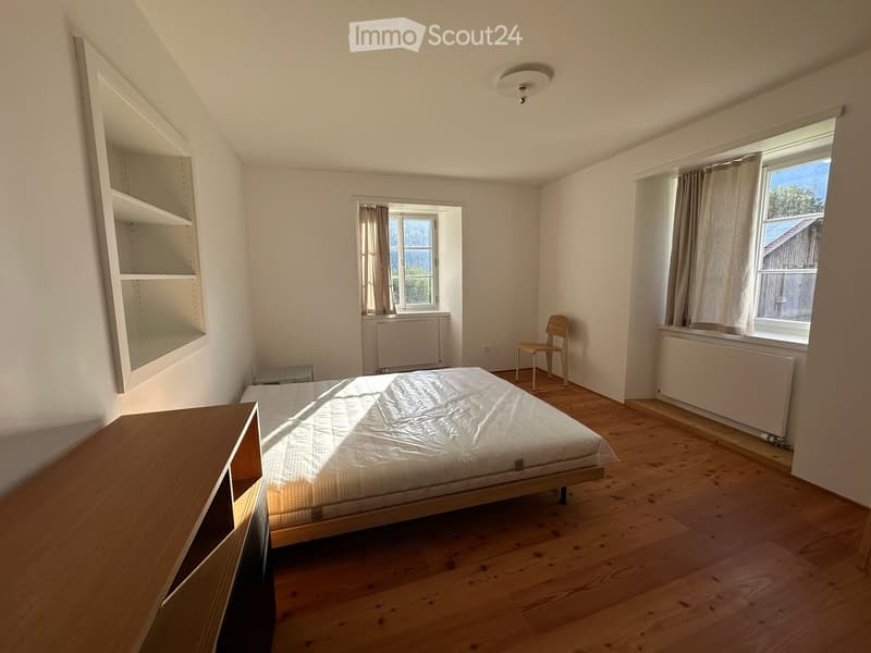 Bright bedroom with wooden flooring, a bed, a chair, a cabinet, and built-in shelves. Natural light streams in through two windows with curtains.
