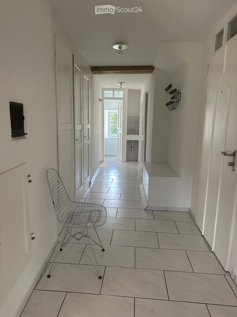 A modern hallway with tiled flooring, white walls, and a single wire chair placed near the left wall. The hallway leads to a small bathroom visible at the end.