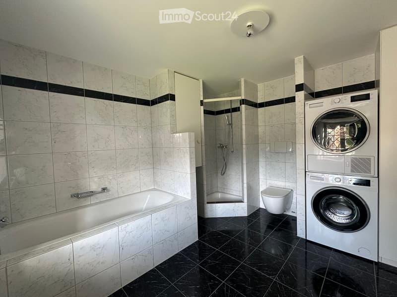 Modern bathroom with marble tiles, bathtub, toilet, shower, and stacked washer/dryer unit.