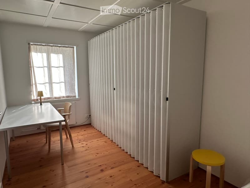 A modern, minimalist room with wooden flooring, white walls, and large windows. Features a white table, a wooden chair, a yellow stool, and a small lamp on the table.