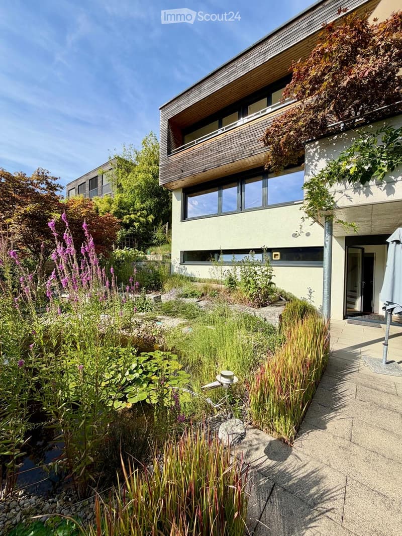Modern two-story house with a balcony, surrounded by a landscaped garden with various plants, shrubs, and a small water feature.