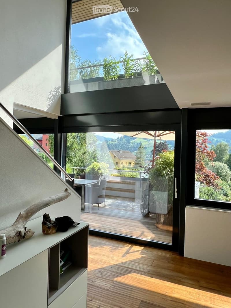 Modern interior with large windows and a balcony overlooking a scenic landscape, featuring a chair, table, and decorative items.