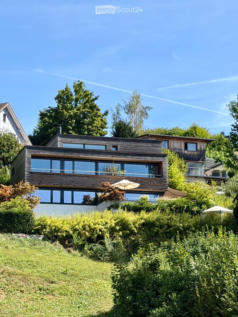 Modern two-story house with large windows and wooden exterior, surrounded by lush greenery and trees.