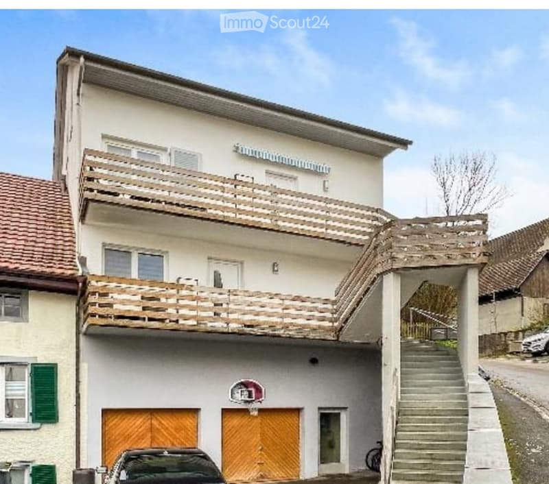 Two-story residential building with white exterior, wooden balconies, garage, and basketball hoop.