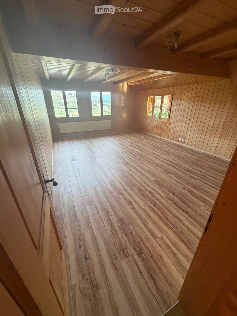 Empty room with wooden flooring, wooden walls, and large windows offering a scenic view. The room features exposed wooden beams on the ceiling and a radiator on one wall.
