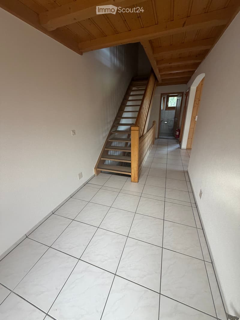 A clean, tiled hallway with a wooden staircase leading upwards, featuring light-colored walls and a visible bathroom entrance.