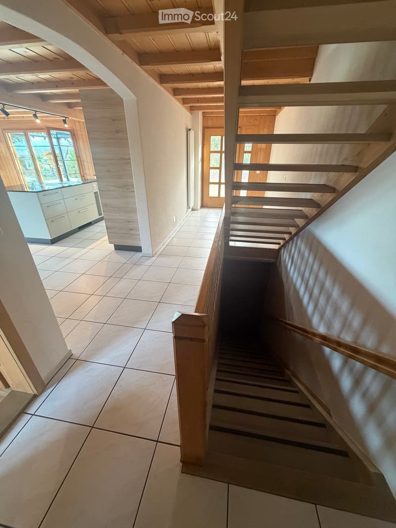 Spacious hallway with wooden staircase, tiled floor, and modern lighting fixtures. The hallway leads to a kitchen area with white cabinets and a large window.