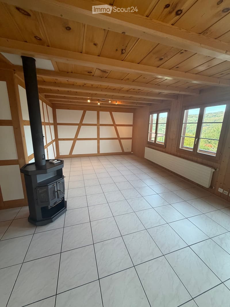 Empty room with exposed wooden beams on the ceiling, tiled floor, large windows with scenic views, and a black wood-burning stove.