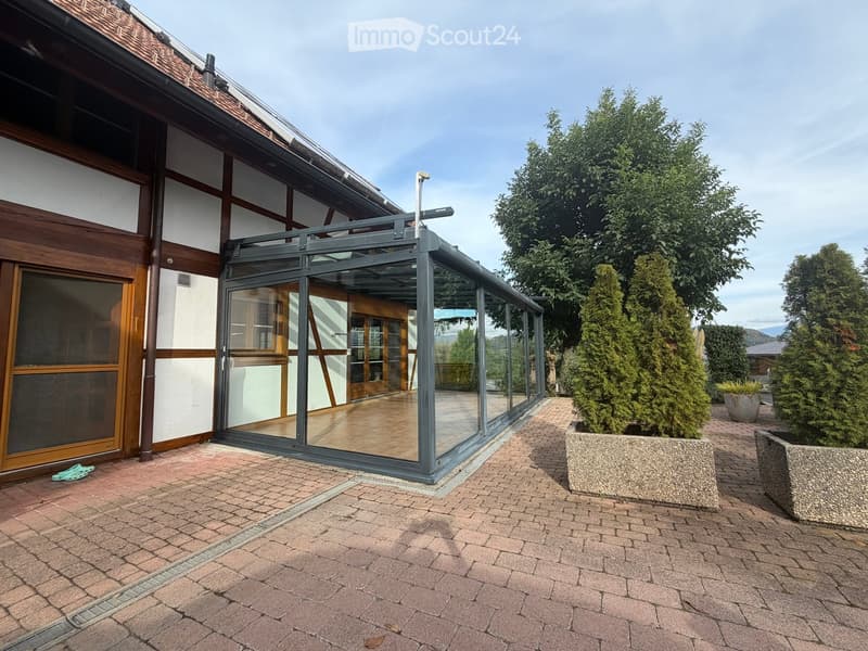 Brick-paved outdoor area with a modern glass-enclosed patio attached to a wooden and white building, surrounded by trees and potted plants.