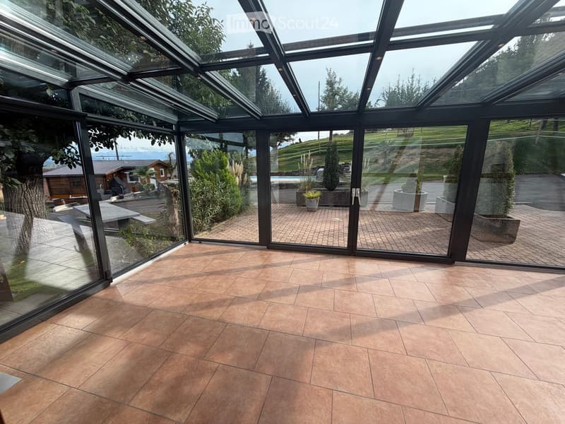 Empty room with tiled floor and large glass walls offering a view of the garden and outdoor area.