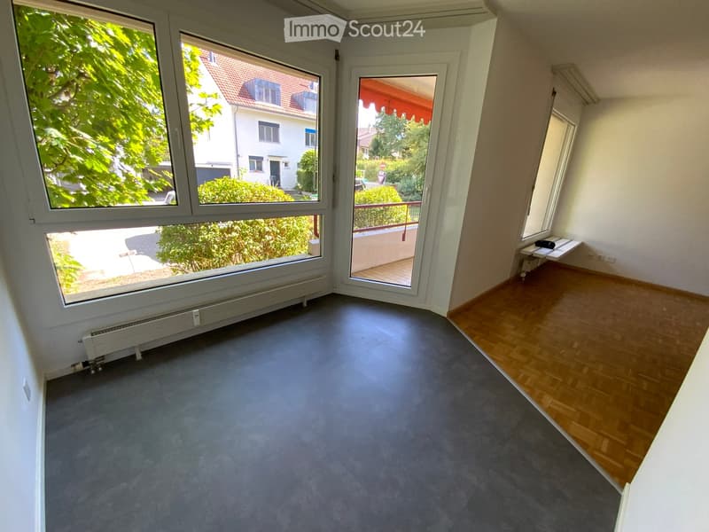 Empty room with grey carpeted floor, wooden flooring in one corner, large windows, and a sliding glass door leading to a balcony.