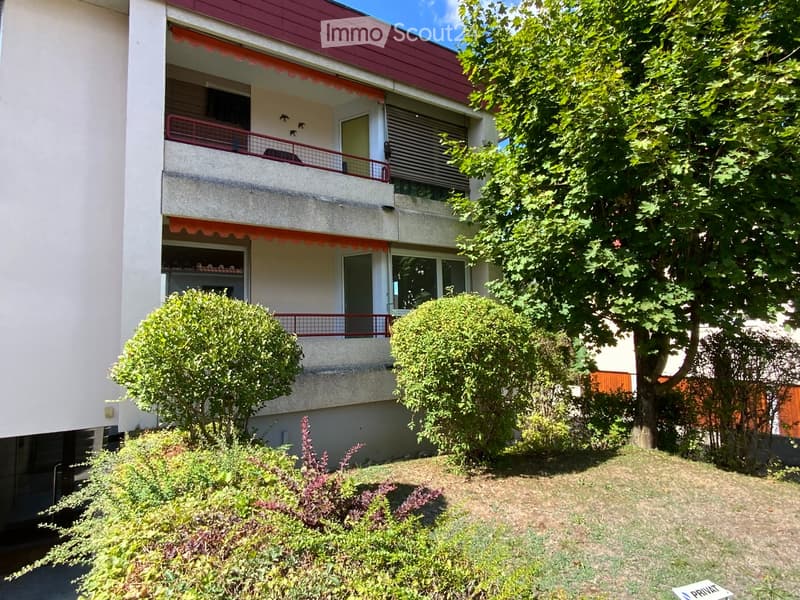 Two-story apartment building with balconies, red roof, and surrounding greenery including trees and shrubs.