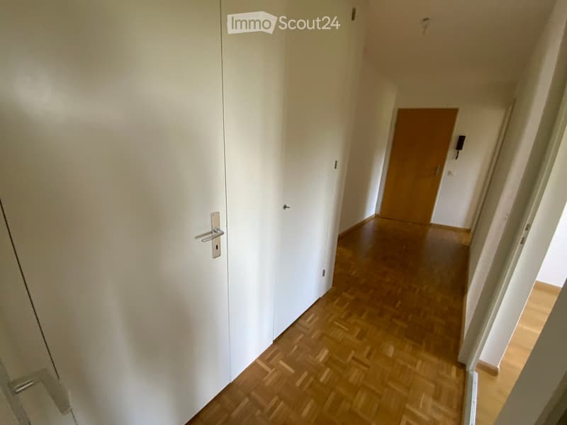 A narrow hallway with wooden flooring and white walls, featuring a closed white door on the left and an open wooden door at the end leading to another room.