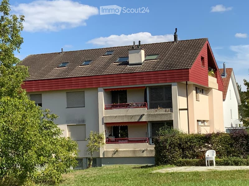 Two-story house with red and beige exterior, multiple windows, balconies, and a small garden area with a white plastic chair.