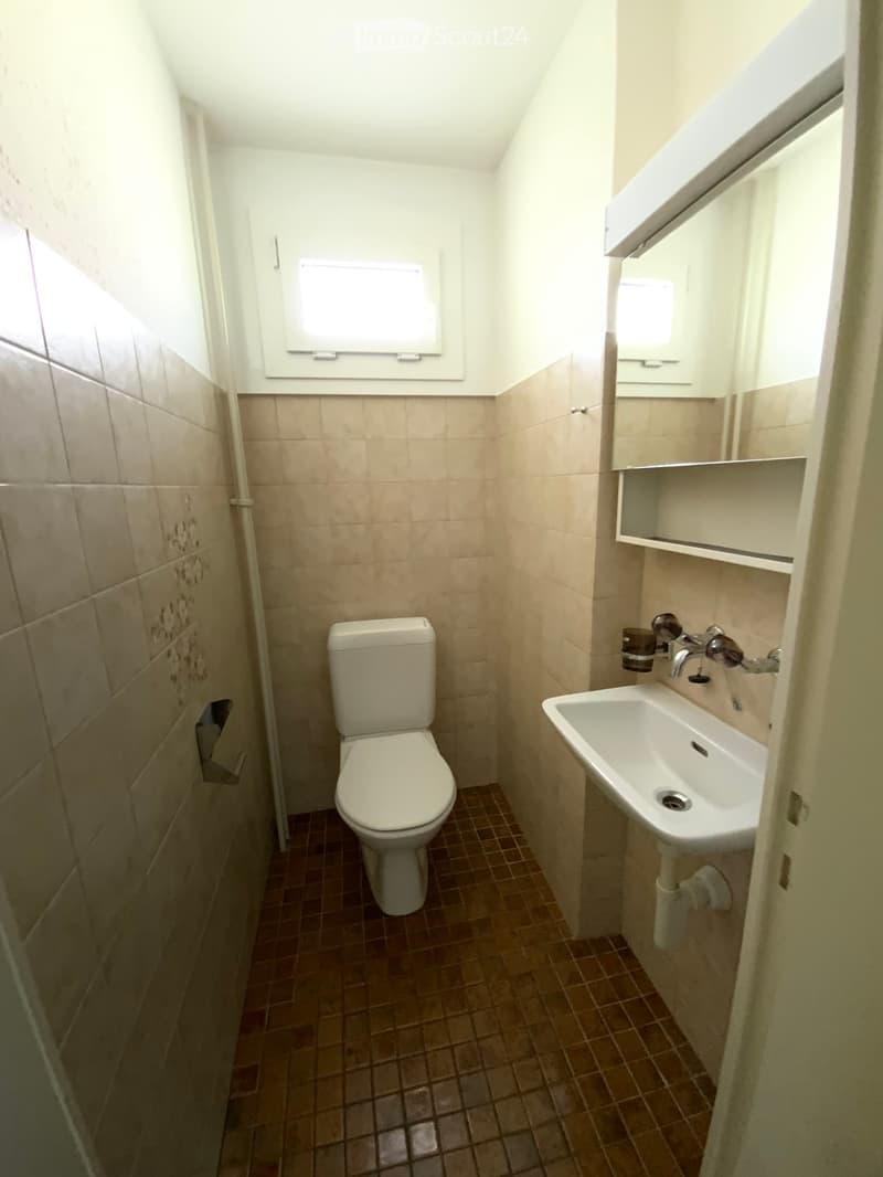Small bathroom with beige tiled walls, brown tiled floor, toilet, sink, faucet, mirror, and cabinet.