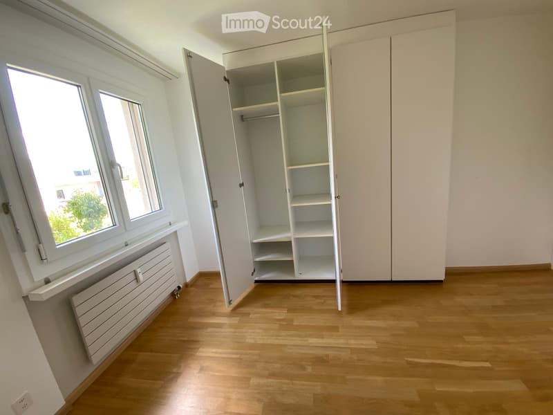 Empty room with wooden flooring, white walls, large window, and built-in closet with empty shelves.