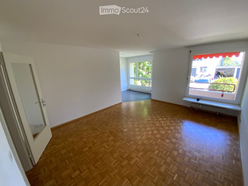 Empty room with wooden herringbone flooring, white walls, large windows, and a door leading to another room.