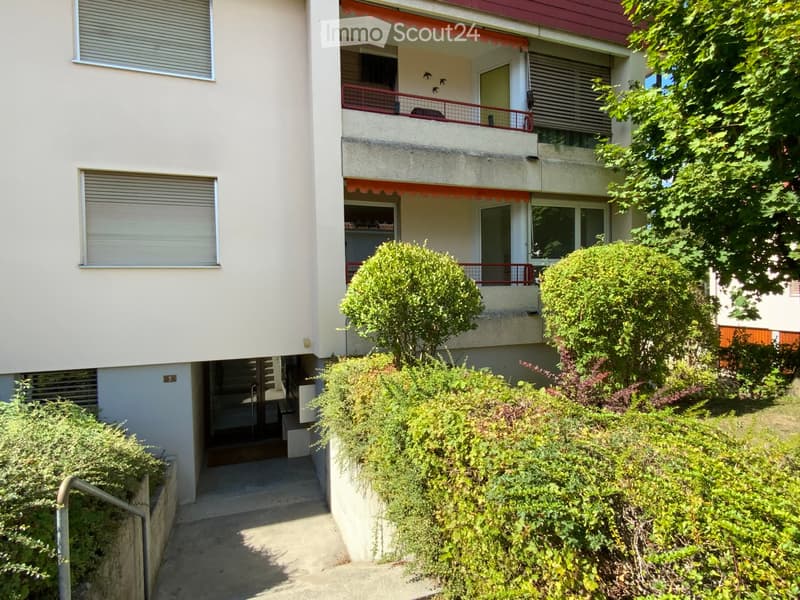 Two-story residential building with balconies, greenery, and a raised ground floor entrance.