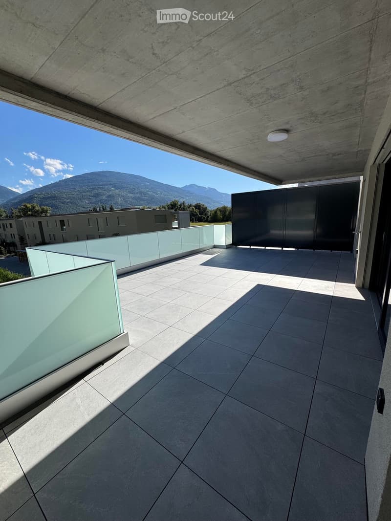 Modern balcony with concrete floor, glass railing, and a view of mountains in the background.