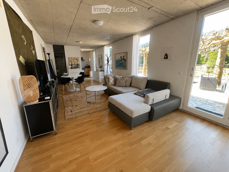 Modern living room with wooden flooring, large windows, a grey sectional sofa, coffee table, dining area with chairs, and a TV on a cabinet.