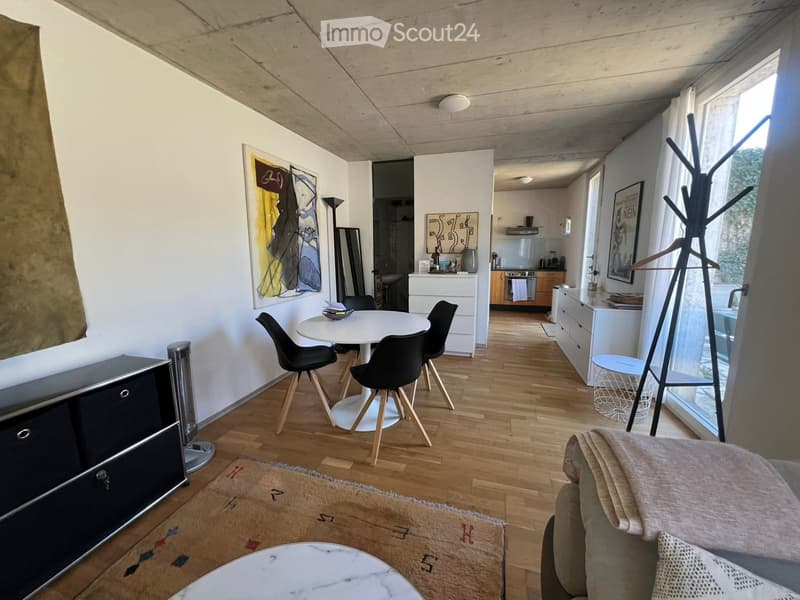 Modern, open-plan living and dining area with wooden flooring, a sofa, coffee table, dining table with chairs, a cabinet, and a coat rack near the entrance.