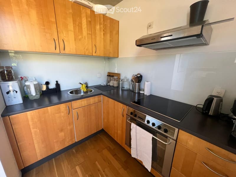 The kitchen features light wooden cabinets, a stainless steel oven, a sink, and various kitchen appliances and utensils on the countertops.