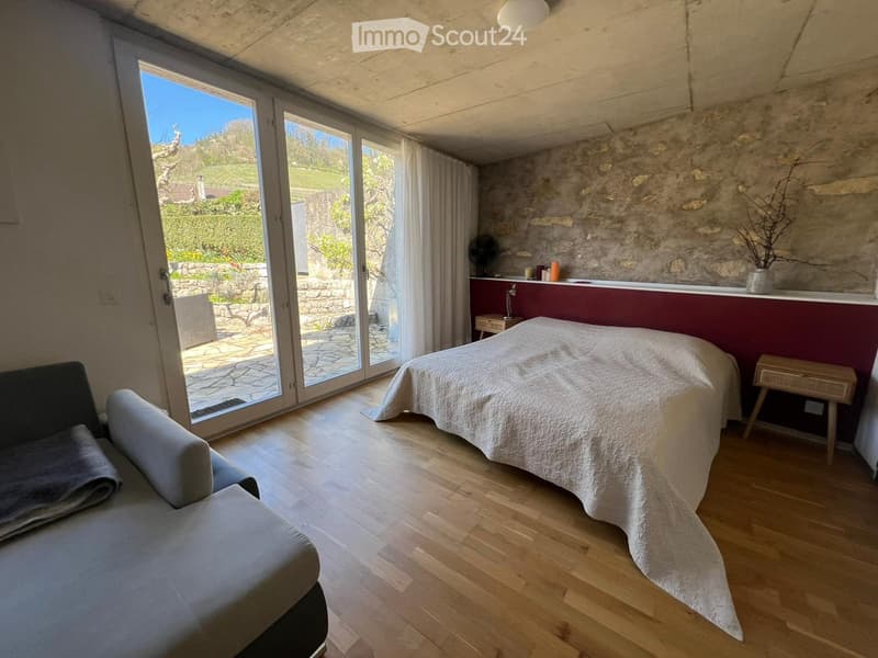 Bright bedroom with wooden flooring, a large bed with white bedding, a grey sofa, a nightstand, and a small table with a fan. Large sliding glass doors open to a garden view.