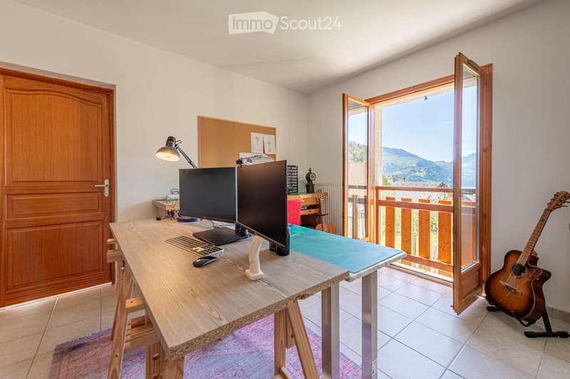 Brightly lit room with a large wooden desk, dual monitors, keyboard, mouse, and a guitar leaning against the wall. A large window offers a scenic mountain view, and a balcony is visible through the open door.