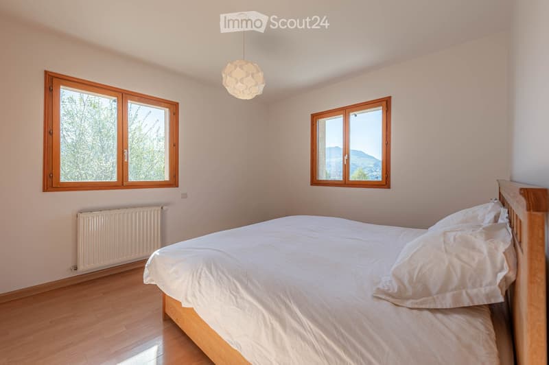 Bright bedroom with wooden flooring, large windows offering scenic views, a neatly made bed, and a radiator below one window.