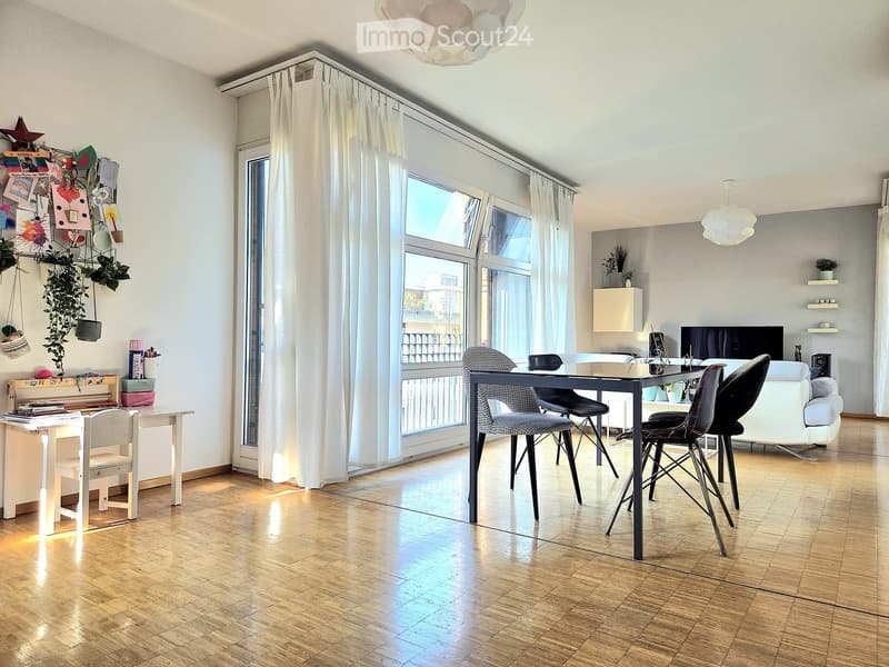 Bright living room with wooden flooring, large windows with white curtains, a sofa, a dining table with chairs, a lamp, and a TV screen.