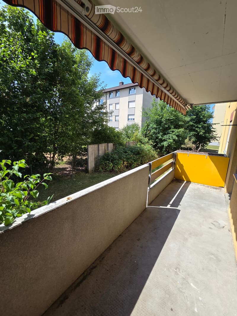 Balcony with concrete floor, metal railing, striped awning, and view of greenery and buildings.