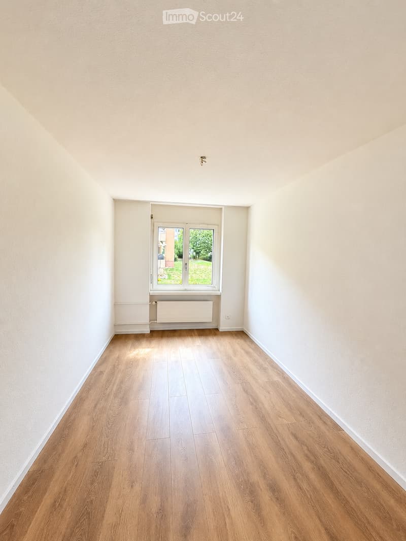 Empty room with light wooden flooring, white walls, and a window with a view of greenery outside. No furniture or equipment is present.