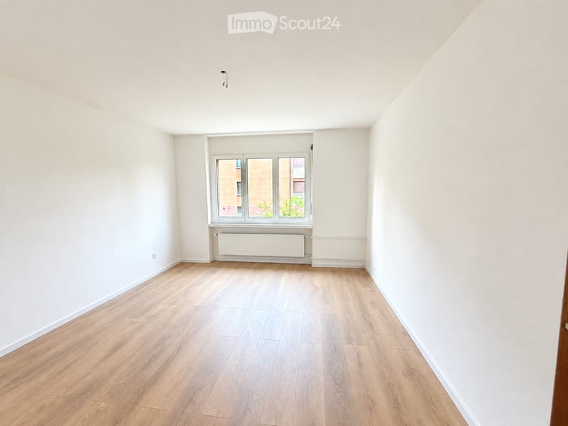 Empty room with light wooden flooring, white walls, and a large window with three panes. A radiator is visible below the window.